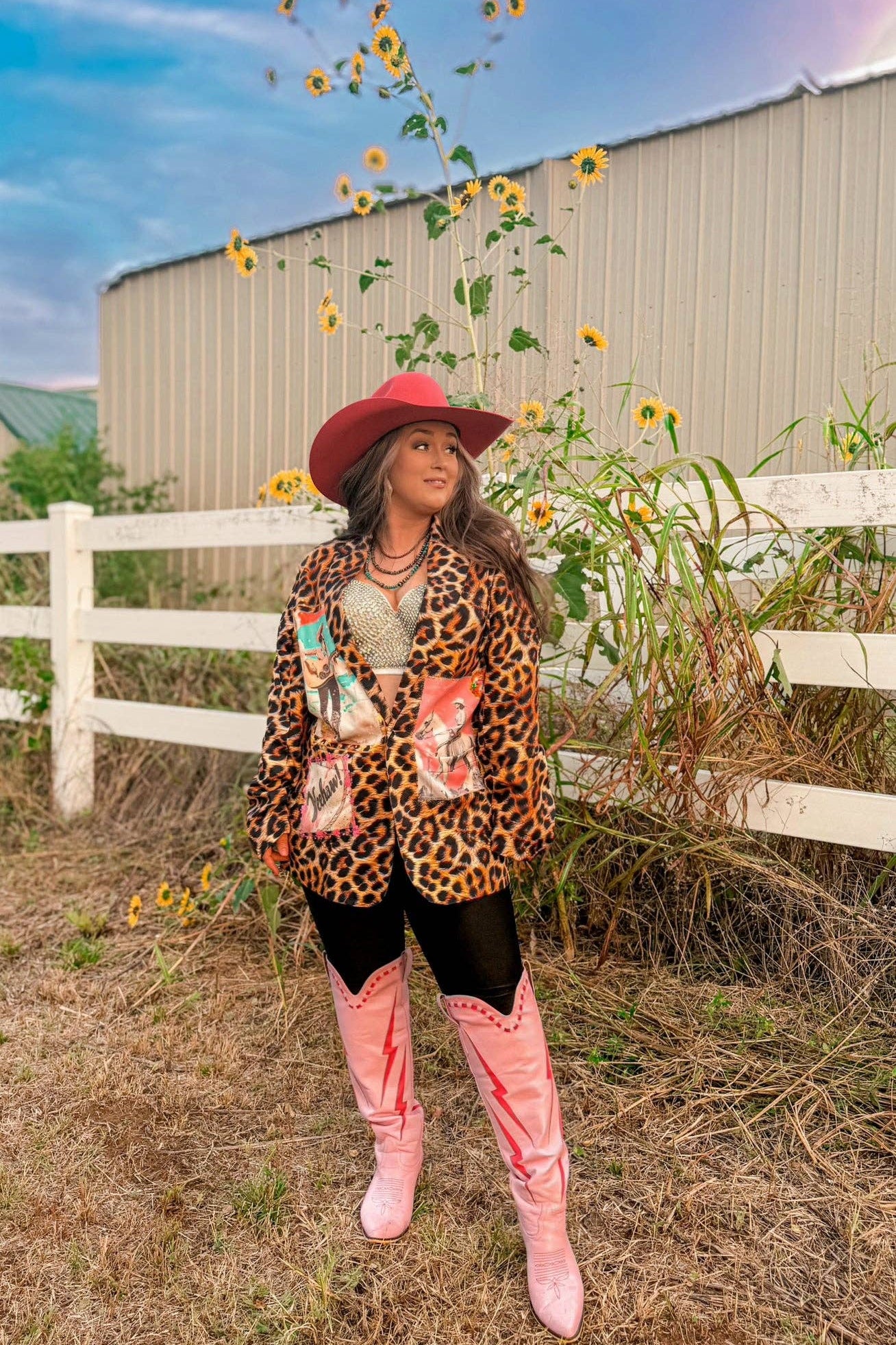 Woman in a leopard print jacket and pink cowboy boots standing in a field with sunflowers and a white fence in the background.