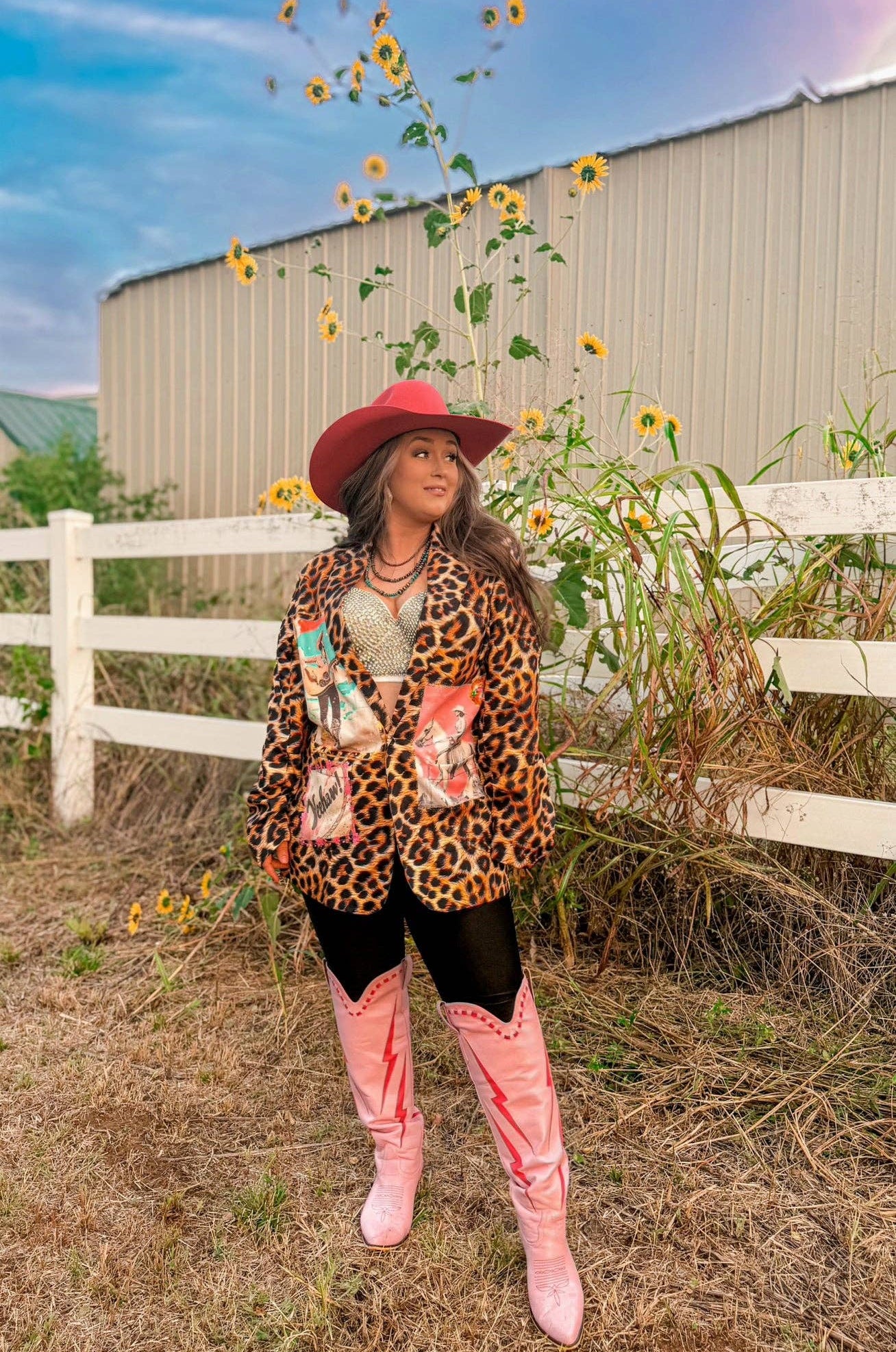 Woman in a leopard print jacket and pink cowboy boots standing in a field with sunflowers and a white fence in the background.