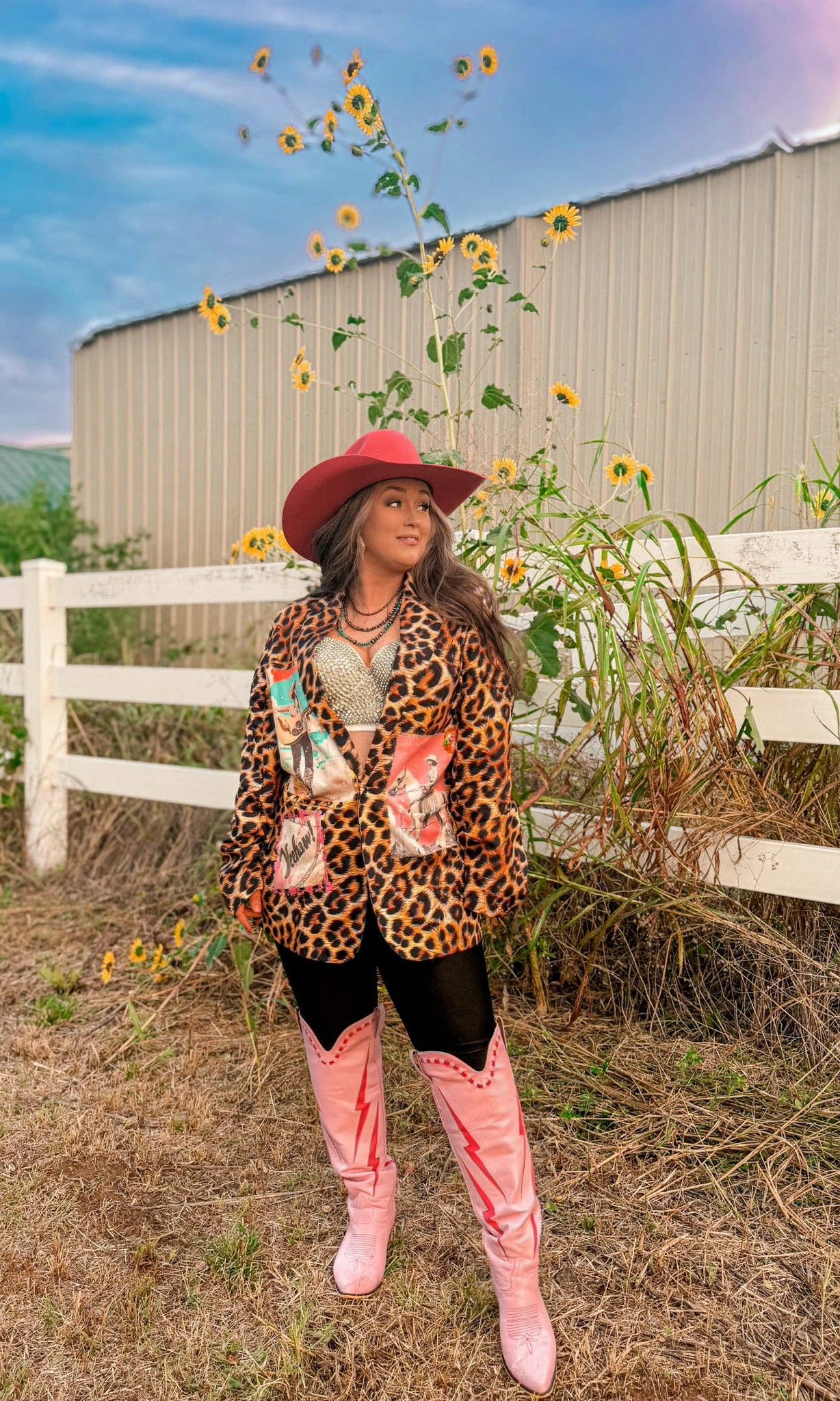 Woman in a leopard print jacket and pink cowboy boots standing in a field with sunflowers and a white fence in the background.