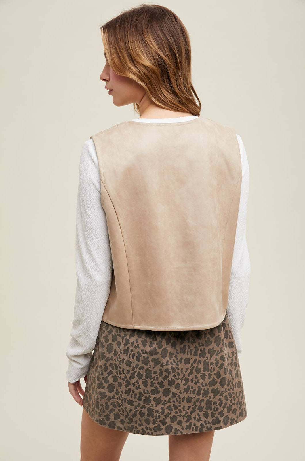 Back of a woman wearing a stone suede snap-up vest over a white shirt, paired with a leopard print mini skirt.