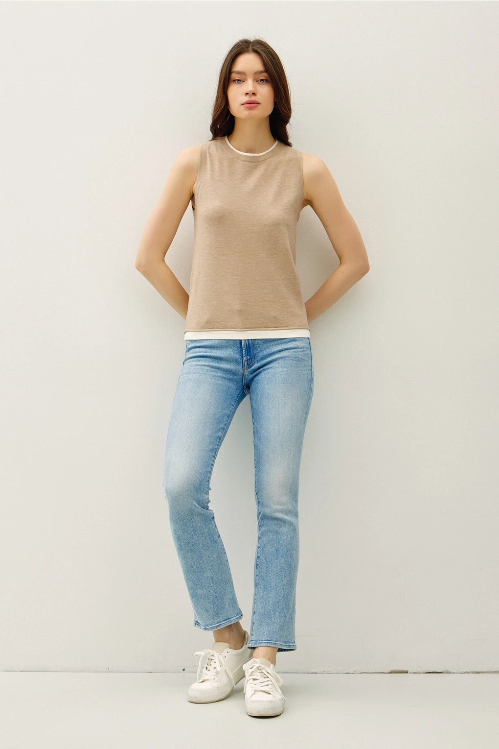 Woman wearing a tan sleeveless top with lighter contrasting band at the bottom of the tops and blue jeans on a plain background