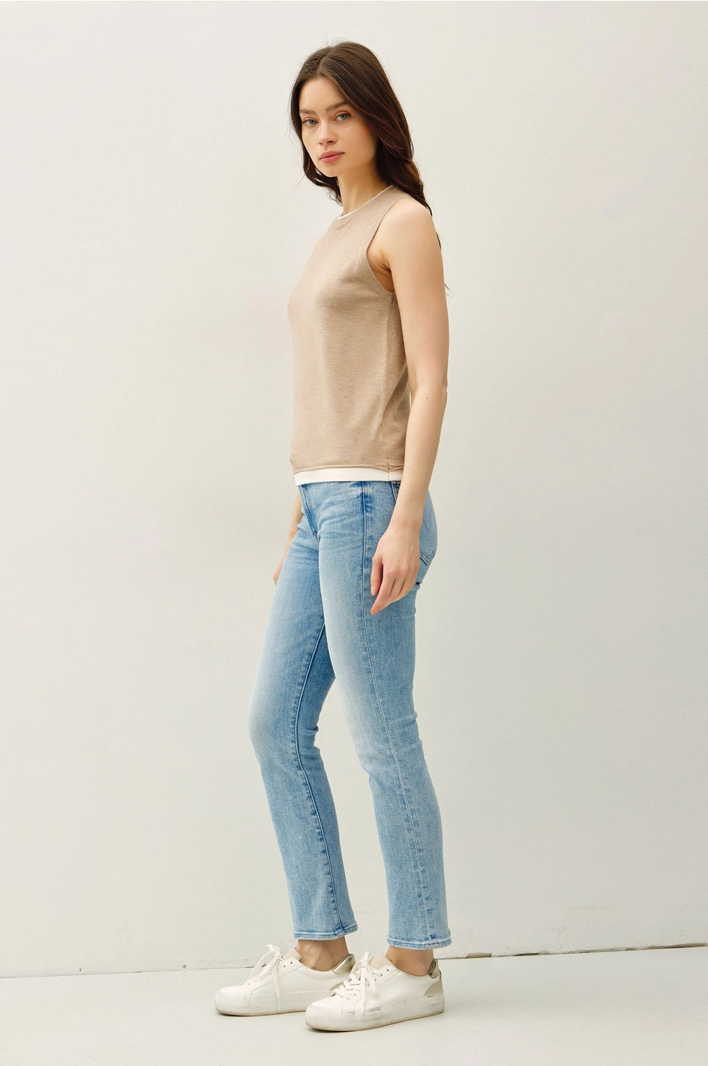 Woman wearing a tan sleeveless top with lighter contrasting band at the bottom of the tops and blue jeans on a plain background