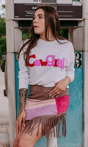 Woman wearing a white sweatshirt with colorful text and a patterned skirt standing next to a payphone.