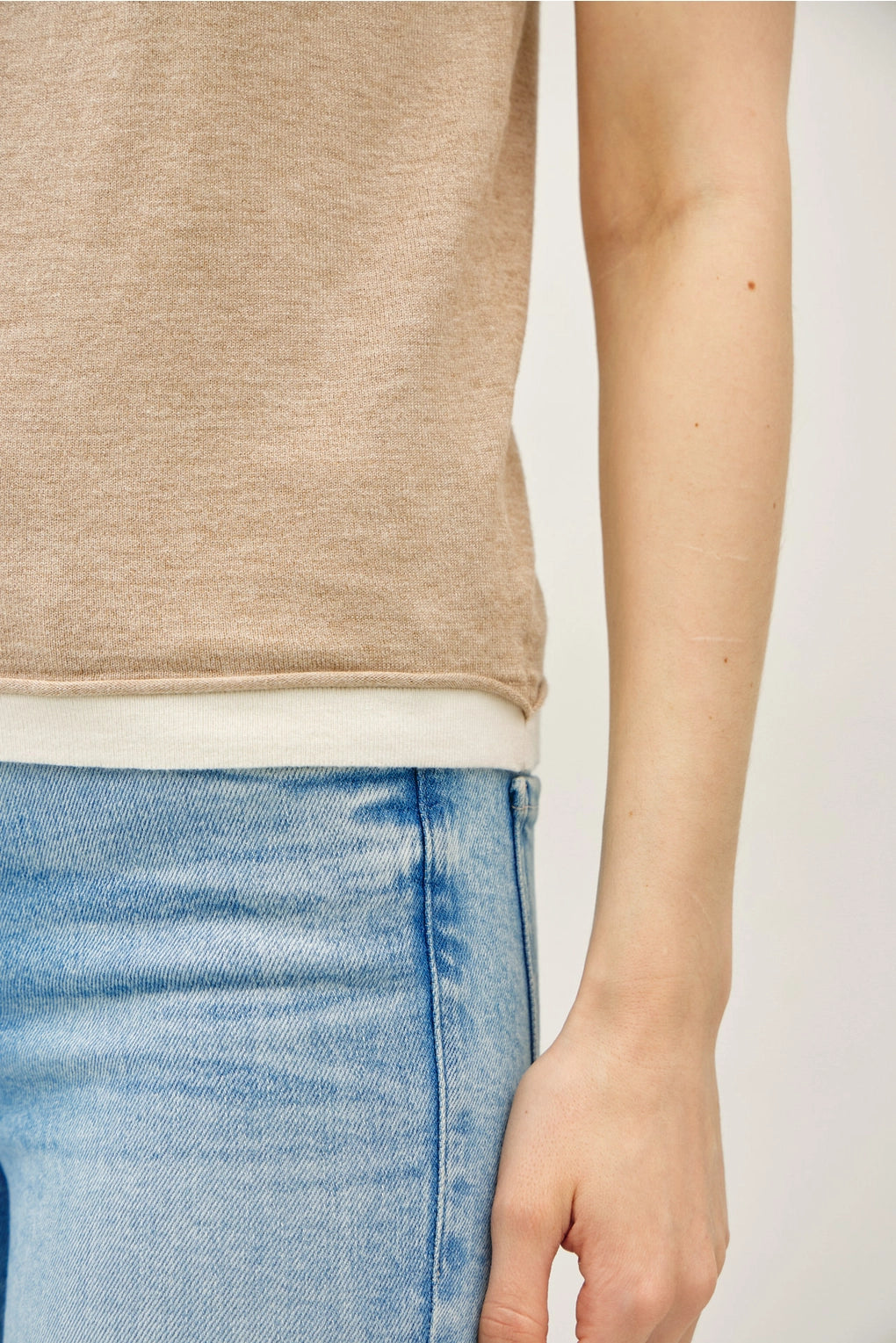 Close up shot of Woman wearing a tan sleeveless top with lighter contrasting band at the bottom of the tops and blue jeans on a plain background