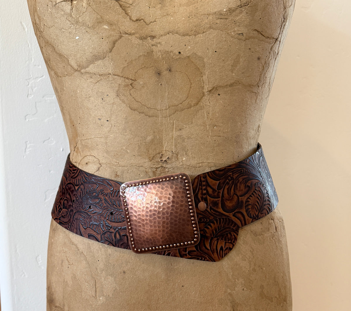 A wide brown tooled leather belt with a hammered copper-toned rectangular buckle, displayed on a vintage tan dress form mannequin.