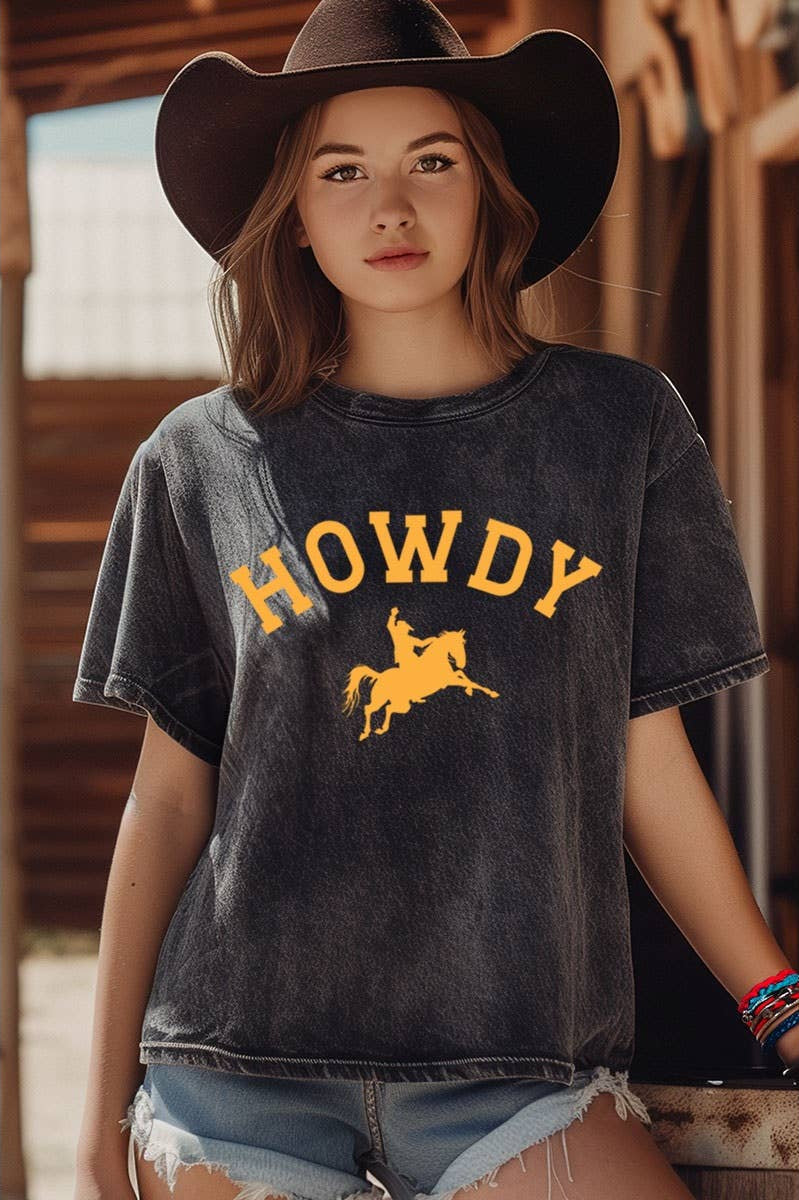 A young woman wearing a dark gray acid-wash t-shirt with a yellow 'HOWDY' cowboy graphic, paired with a black cowboy hat and denim shorts, posed in front of a rustic wooden barn background.