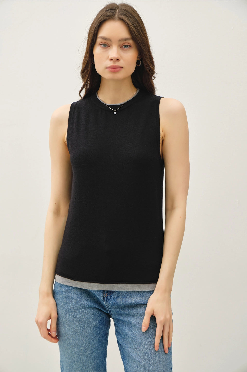 Woman wearing a black sleeveless top with lighter contrasting band at the bottom of the tops and blue jeans on a plain background