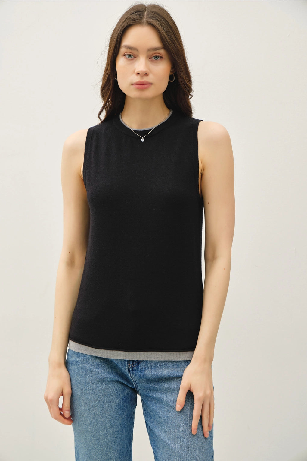 Woman wearing a black sleeveless top with lighter contrasting band at the bottom of the tops and blue jeans on a plain background