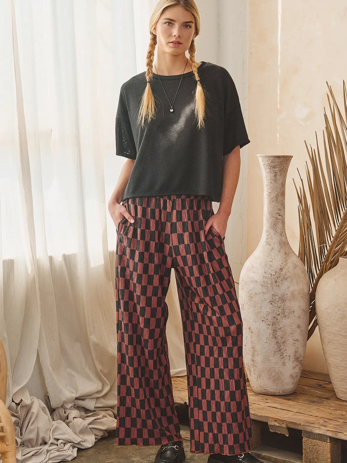 A woman stands indoors wearing a dark charcoal short-sleeve top and wide-leg trousers with a red and black checkerboard pattern. She is posed in a rustic room with sheer curtains, large ceramic vases, and dried palm leaves.