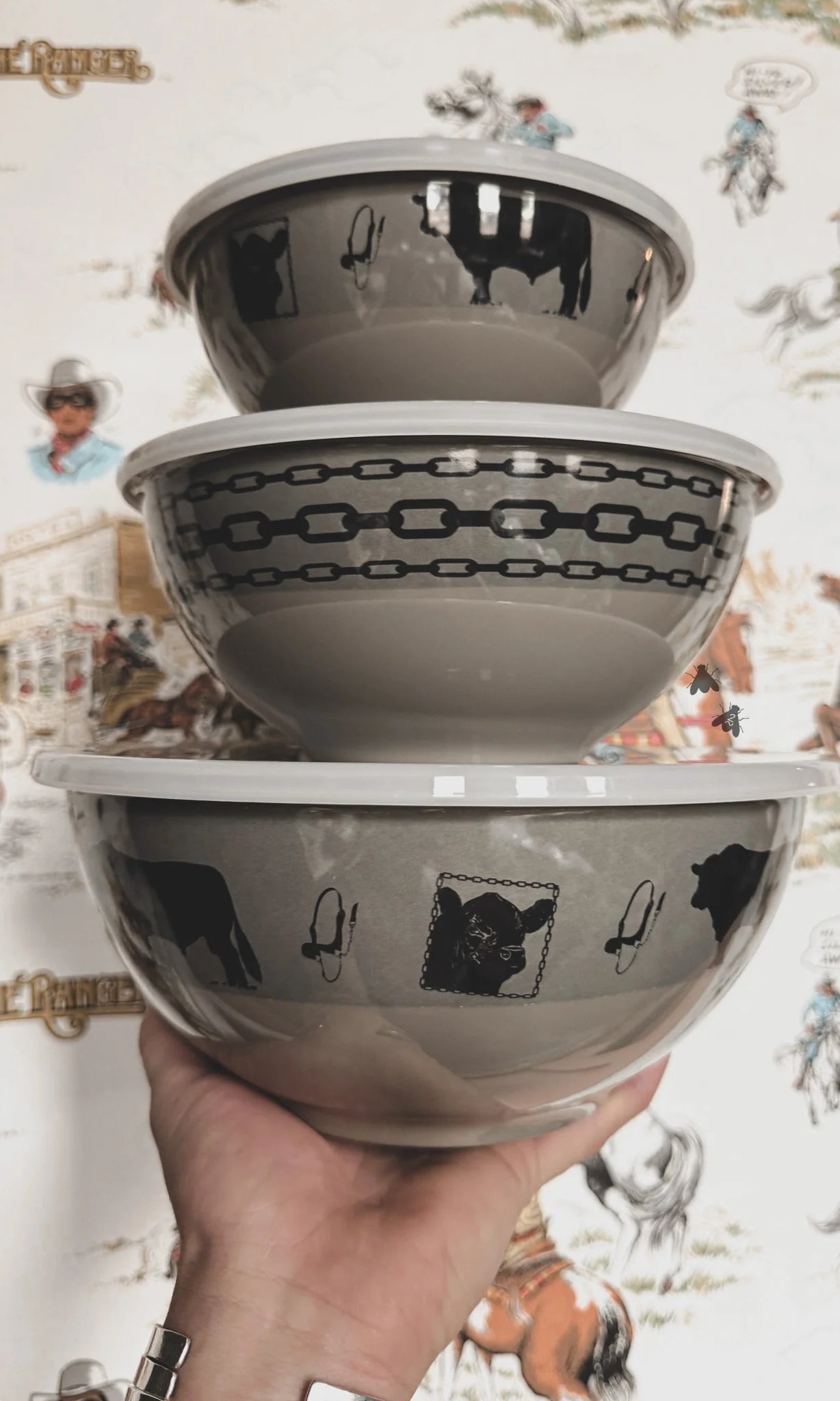 Stack of three melamine bowls with lids and black angus cow patterns held against a wallpapered background.