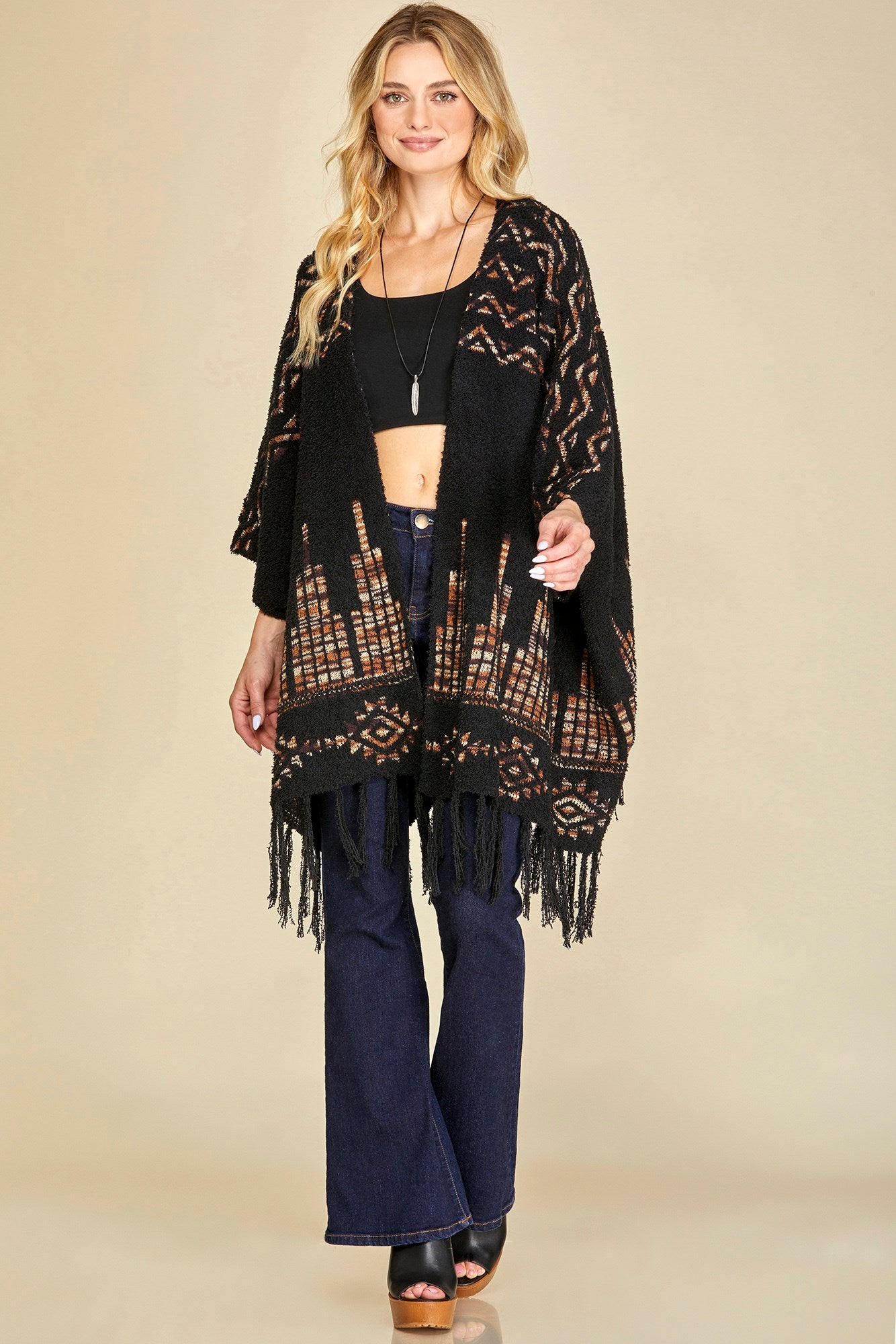 A woman wearing a black and brown aztec patterned poncho with fringe hem, paired with blue jeans and black shoes.