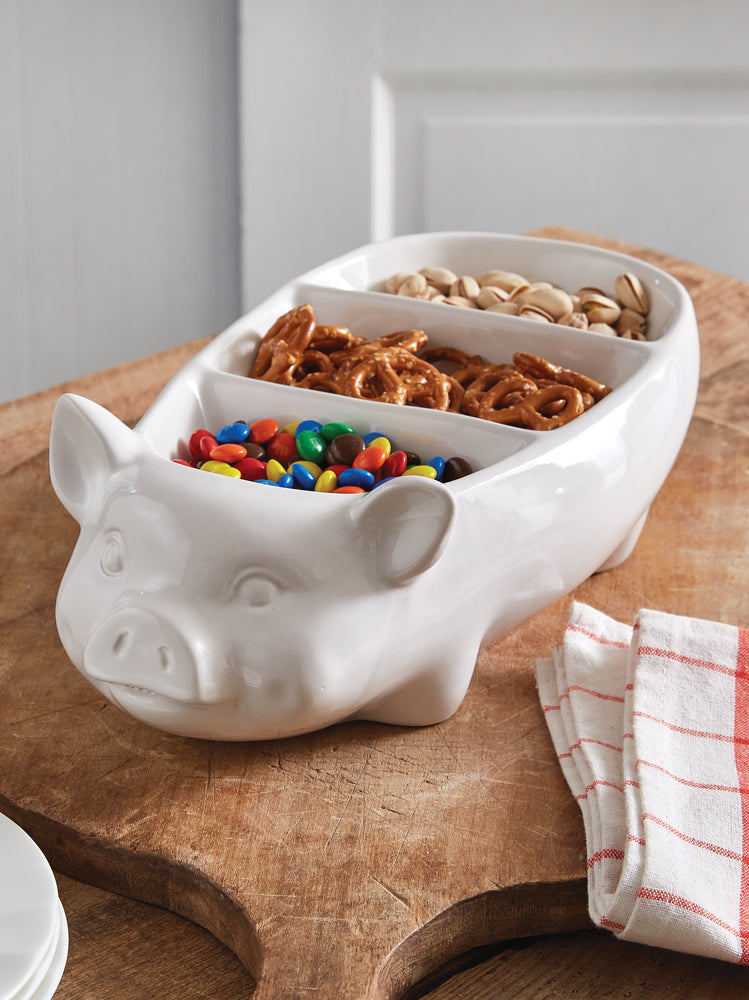 White pig-shaped bowl with compartments filled with snacks on a wooden surface.