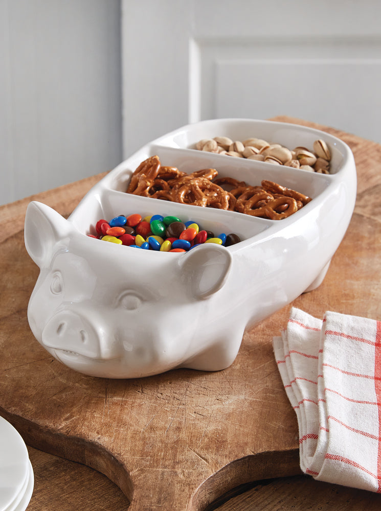 White pig-shaped bowl with compartments filled with snacks on a wooden surface.