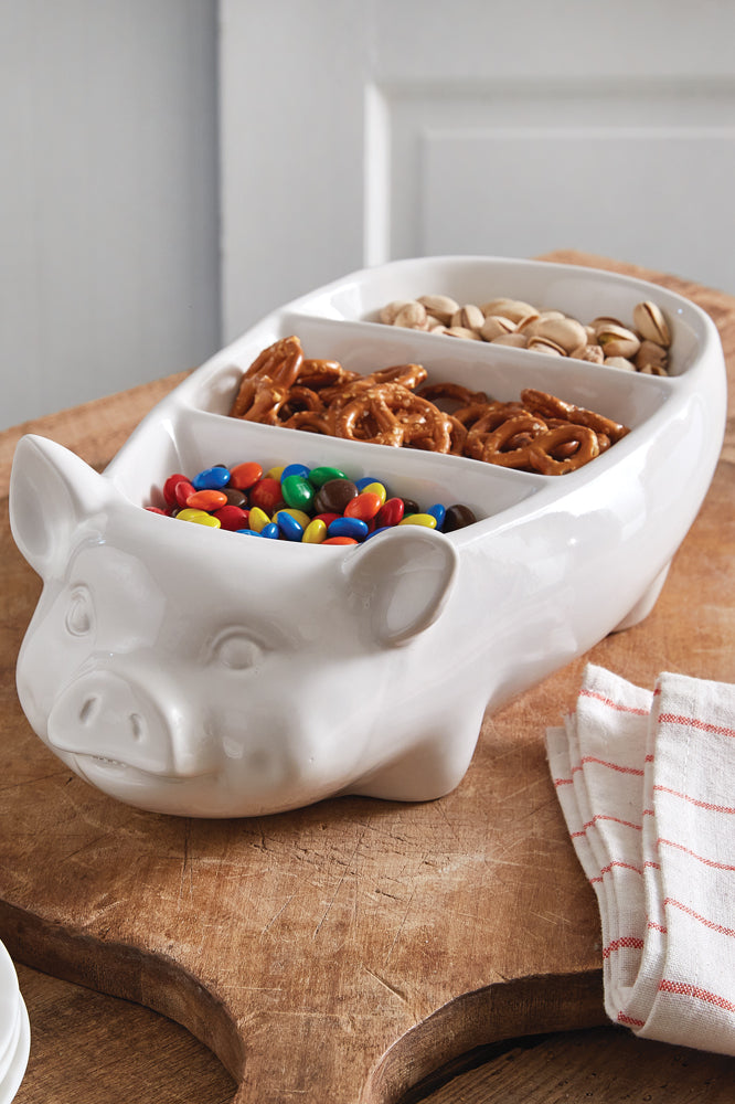 White pig-shaped bowl with compartments filled with snacks on a wooden surface.