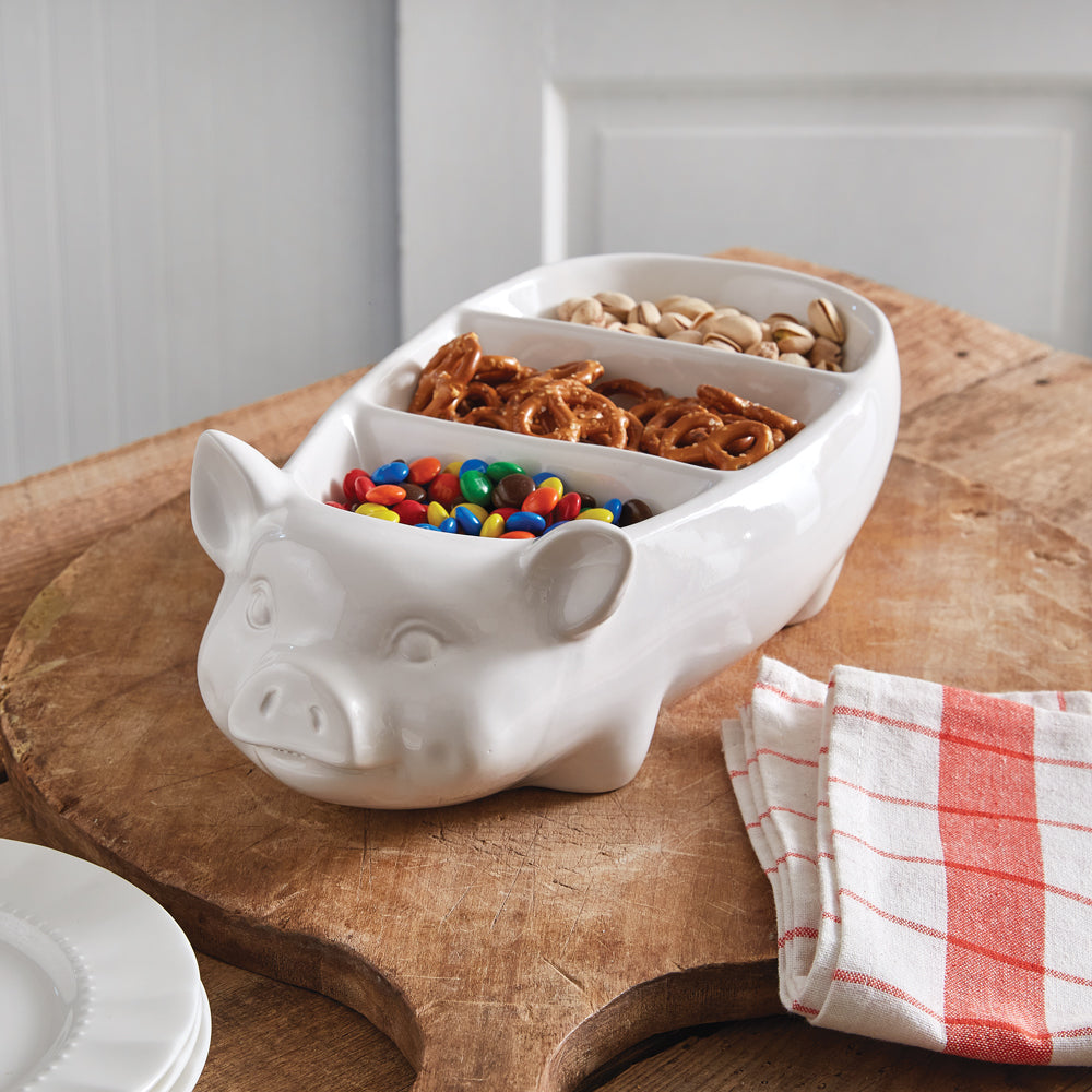White pig-shaped bowl with compartments filled with snacks on a wooden surface.