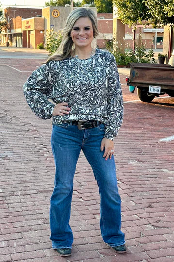 Woman wearing a patterned top and blue jeans standing on a brick sidewalk.