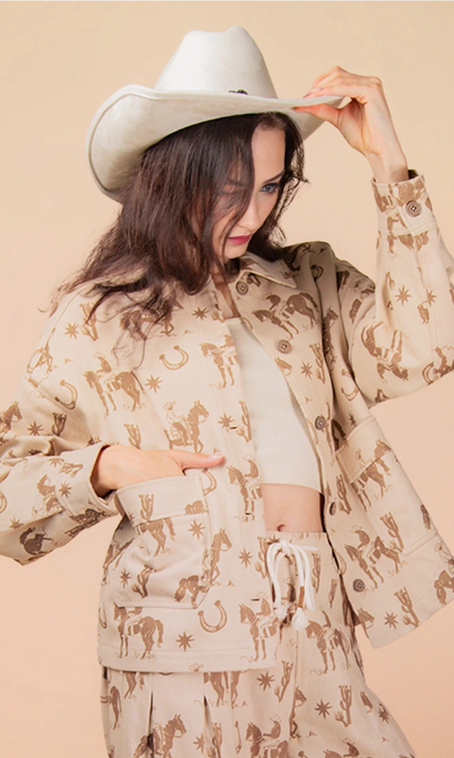 Woman wearing a beige cowboy and horseshoe patterned Shacket and white hat against a beige background