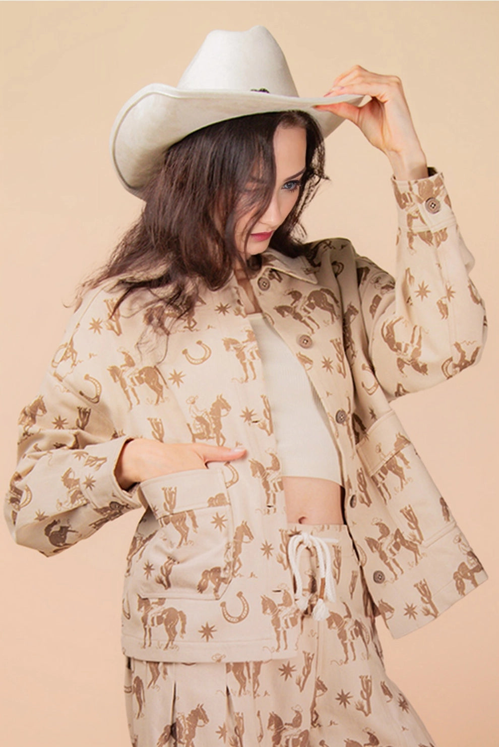 Woman wearing a beige cowboy and horseshoe patterned Shacket and white hat against a beige background