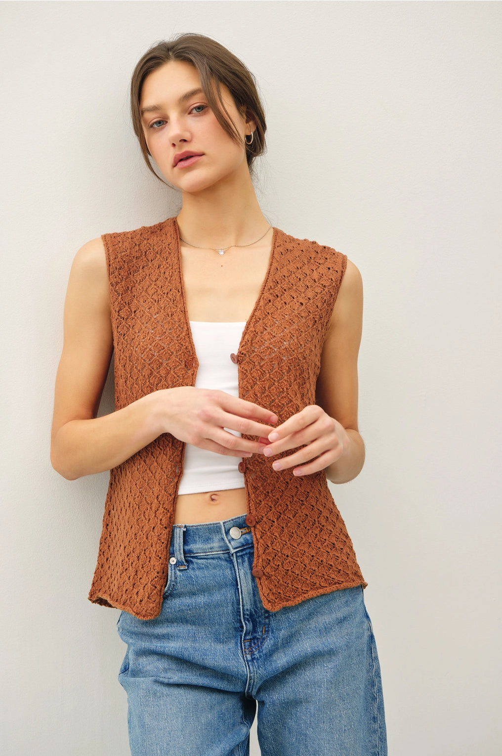 Woman wearing a brown knitted vest over a white top and blue jeans against a plain background