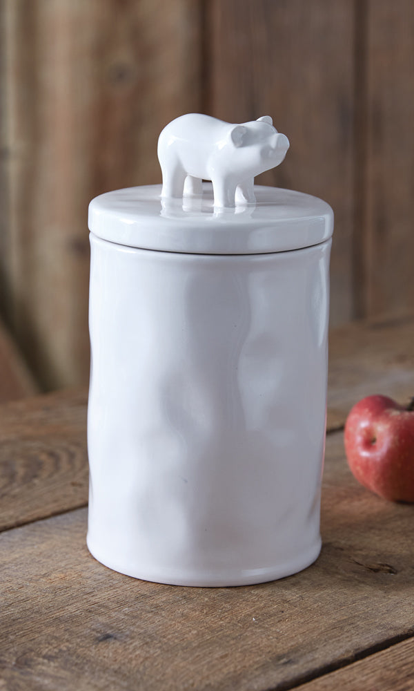 White ceramic jar with a piglet on top, placed on a wooden surface with an apple beside it.
