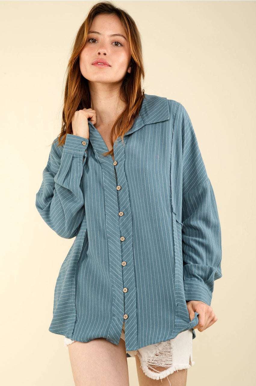 Woman wearing oversized dusty-blue pinstripe button-up collared shirt with drop shoulders and long sleeves; front three-quarter view.”