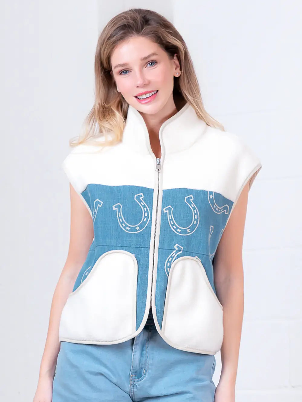 Model wearing indigo denim vest with white horseshoe print and faux-fur collar, zipped front, hands in pockets.”