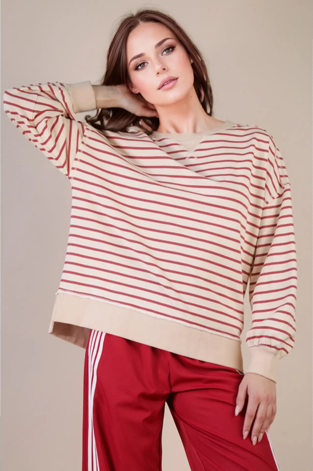 Front view: “Woman wearing an oversized beige and brick striped knit top with drop shoulders, V-stitch crew neckline, and ribbed hem—styled with red track pants.”