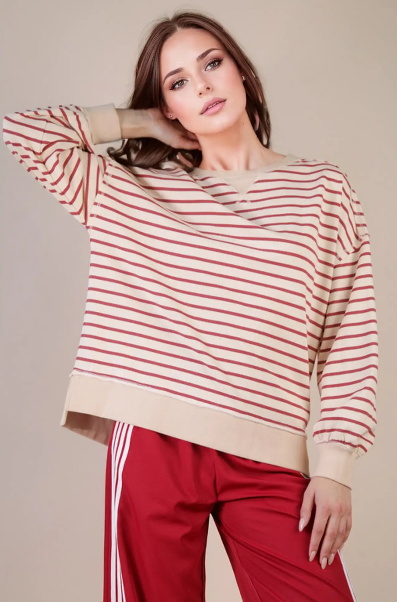 Front view: “Woman wearing an oversized beige and brick striped knit top with drop shoulders, V-stitch crew neckline, and ribbed hem—styled with red track pants.”