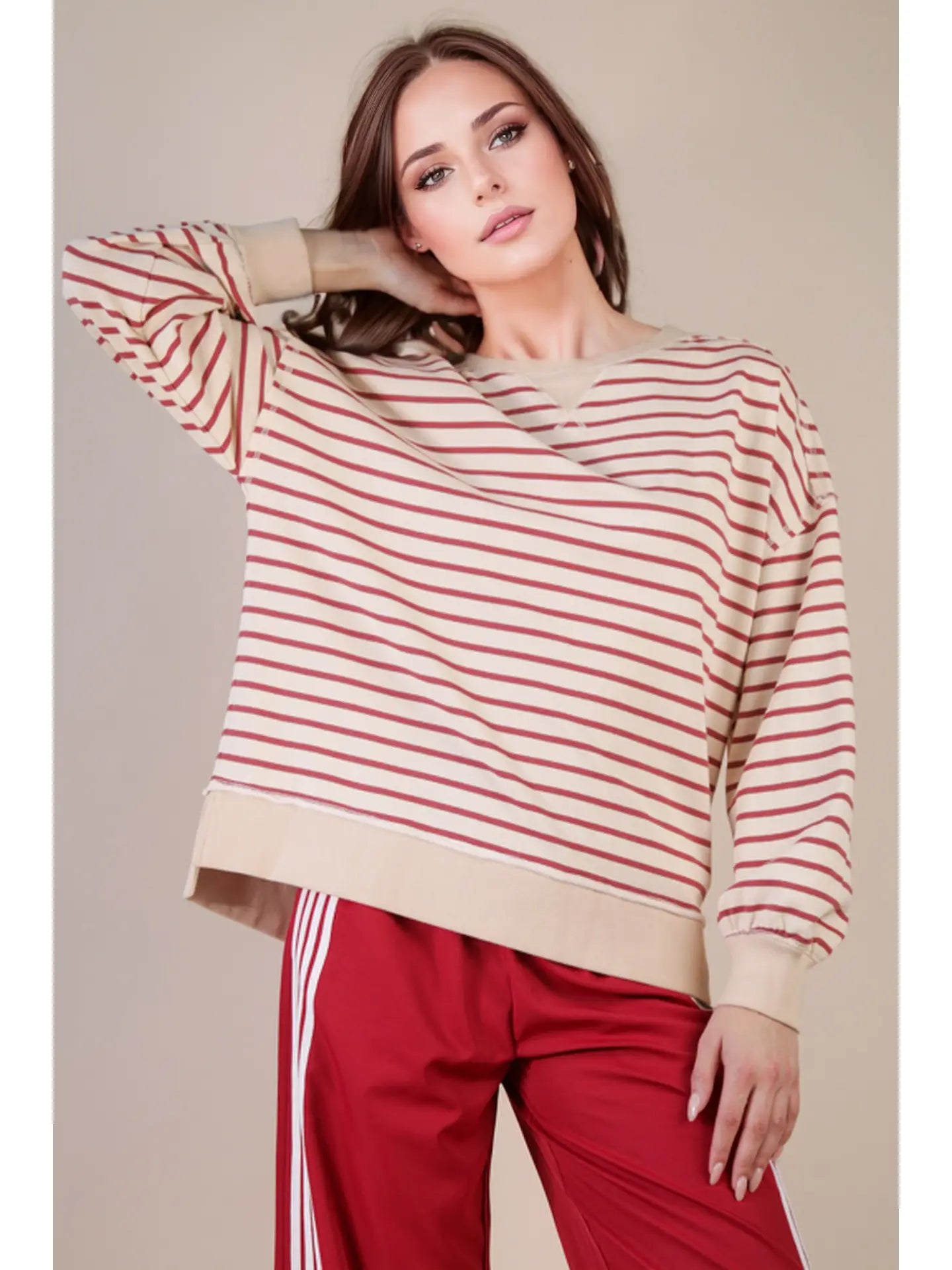 Front view: “Woman wearing an oversized beige and brick striped knit top with drop shoulders, V-stitch crew neckline, and ribbed hem—styled with red track pants.”