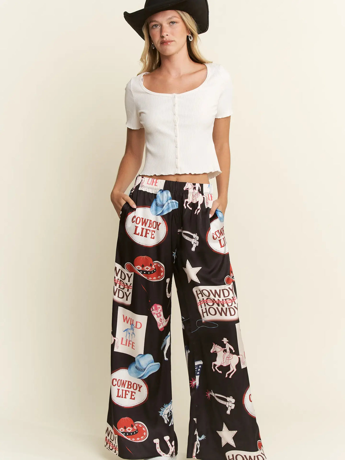 A model wearing a white short-sleeve cropped top and black wide-leg pants with a colorful 'Cowboy Life' graphic print, complete with a black cowboy hat, against a plain cream background.