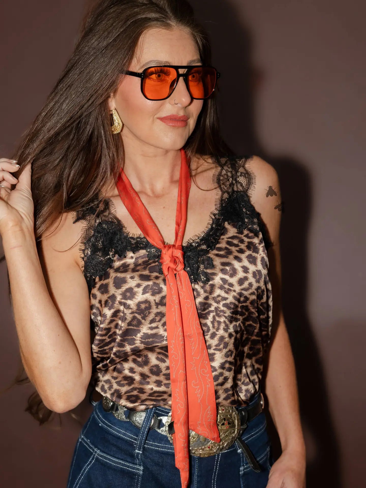 Woman wearing a leopard print top with an orange scarf and sunglasses against a plain background
