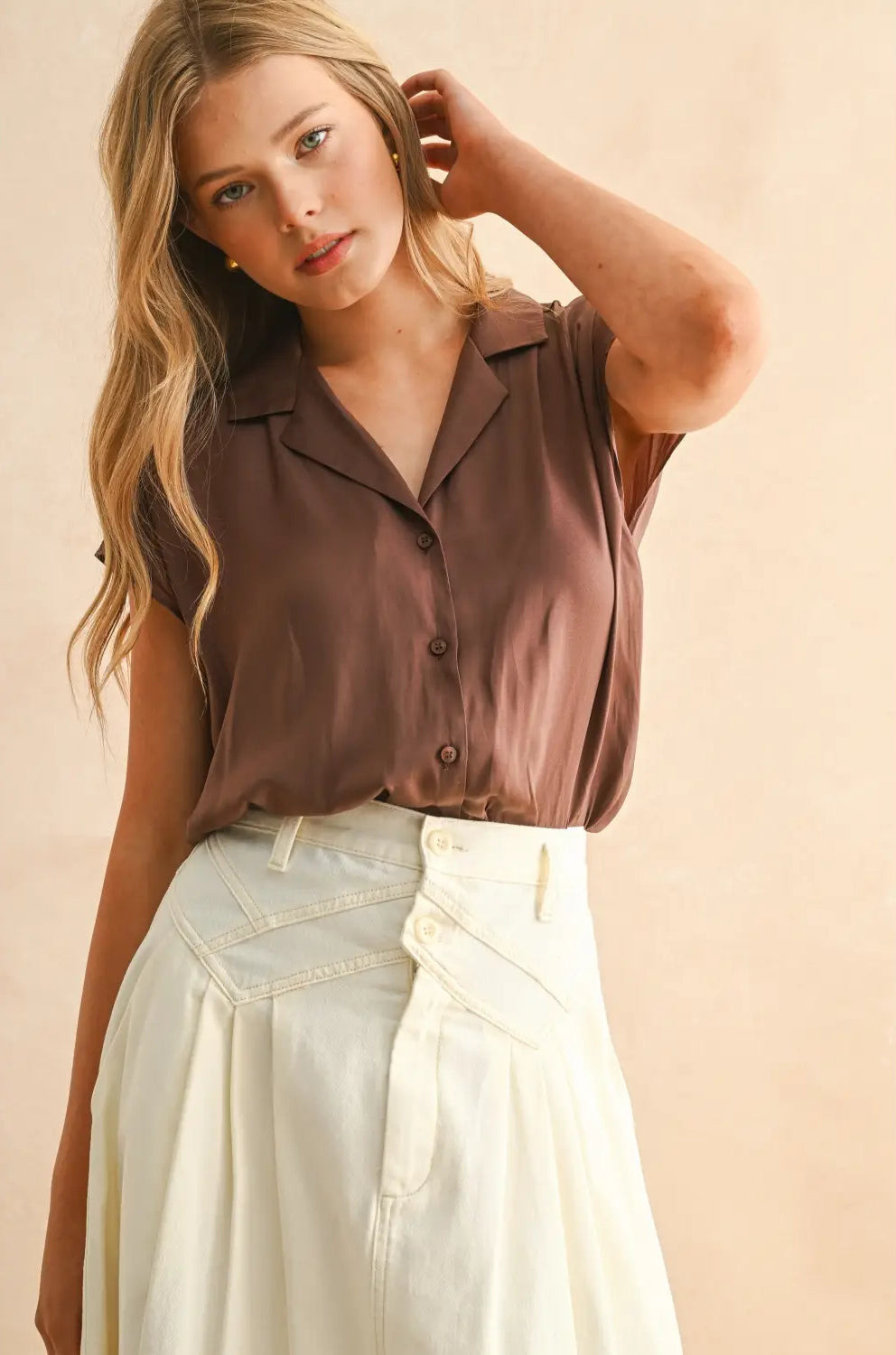 Woman wearing a brown sleeveless blouse and light-colored pants against a beige background