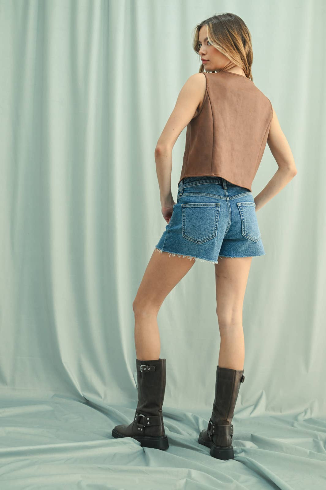 Back of Aa woman wearing a camel suede button-up vest over a white shirt, paired with Denim Shorts