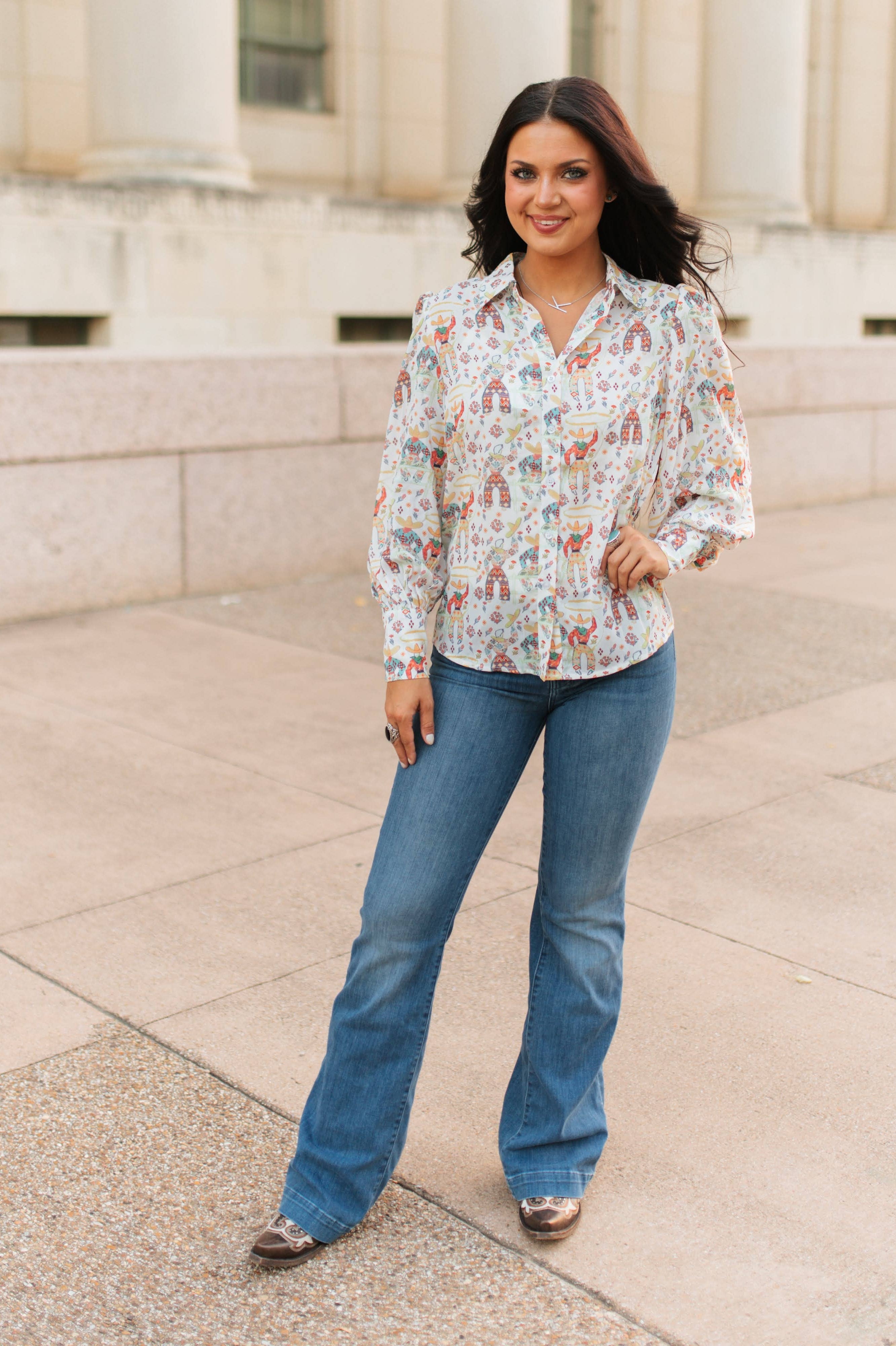 Woman wearing a patterned blouse and blue jeans standing on a stone pavement.