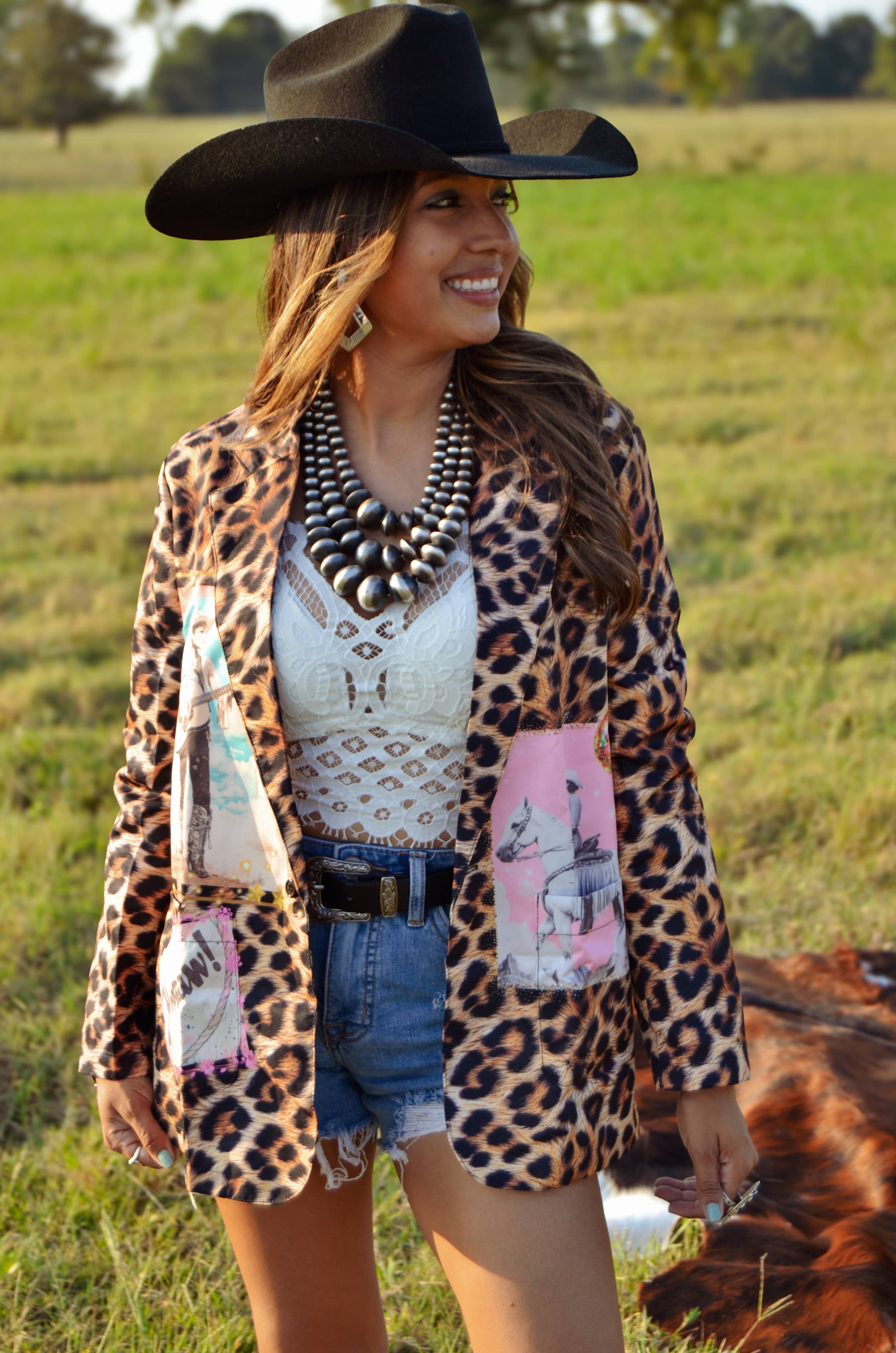 Woman in a leopard print cardigan and cowboy hat standing in a field with a cow.