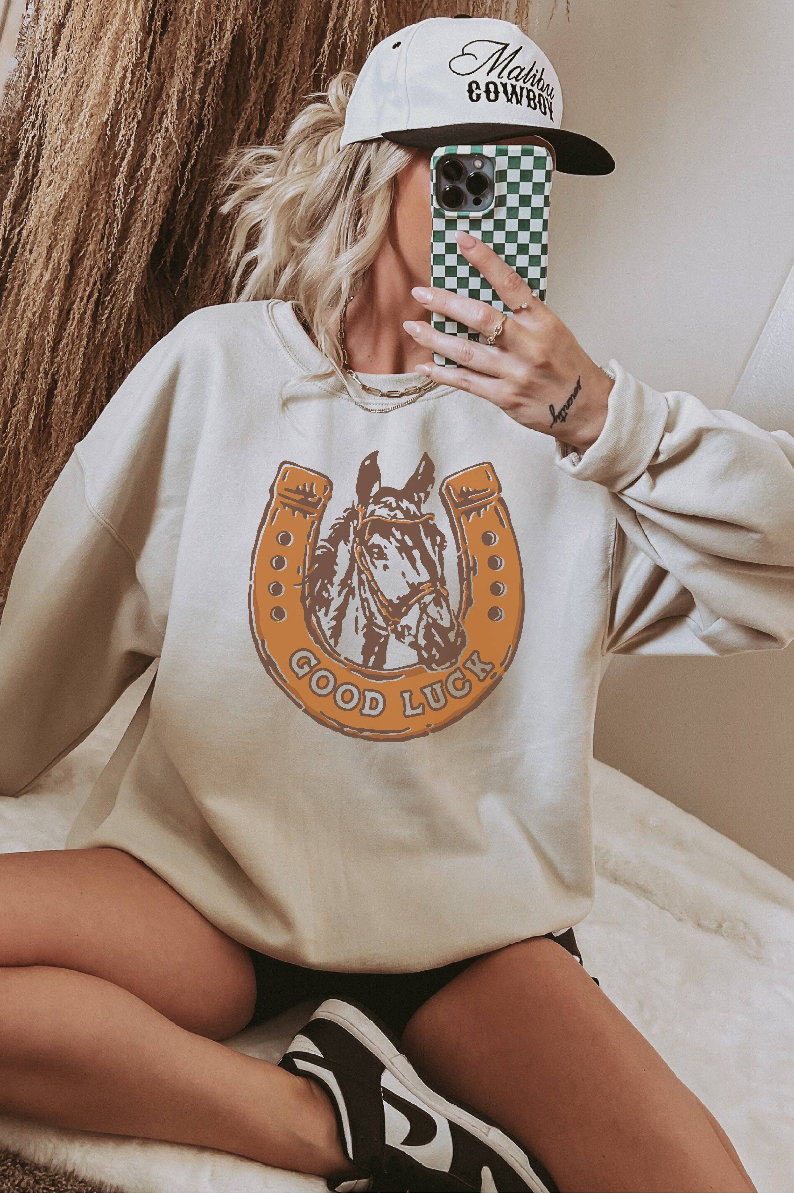 Person wearing a beige sweatshirt with a horse graphic and text, holding a phone with a checkered case.