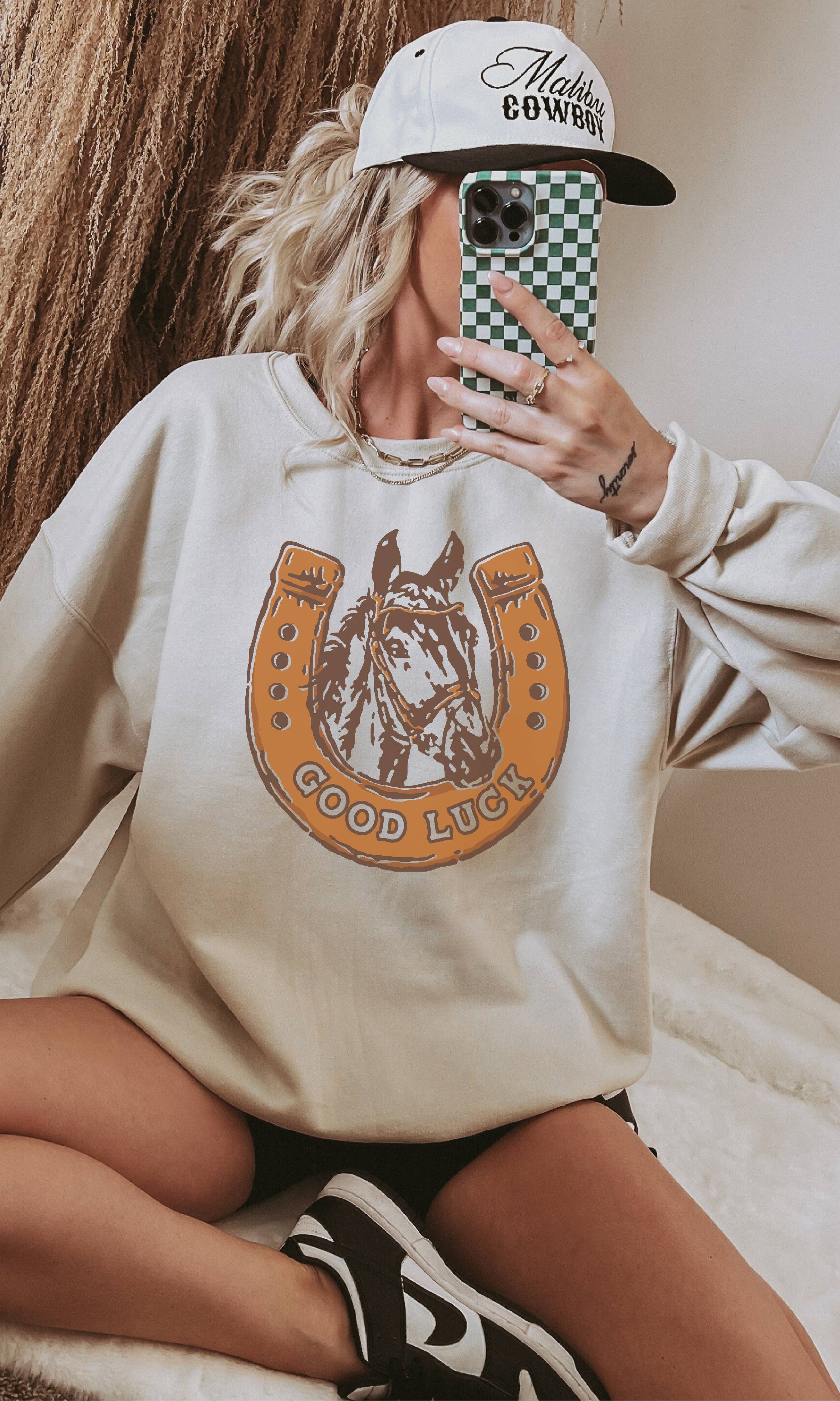 Person wearing a beige sweatshirt with a horse graphic and text, holding a phone with a checkered case.