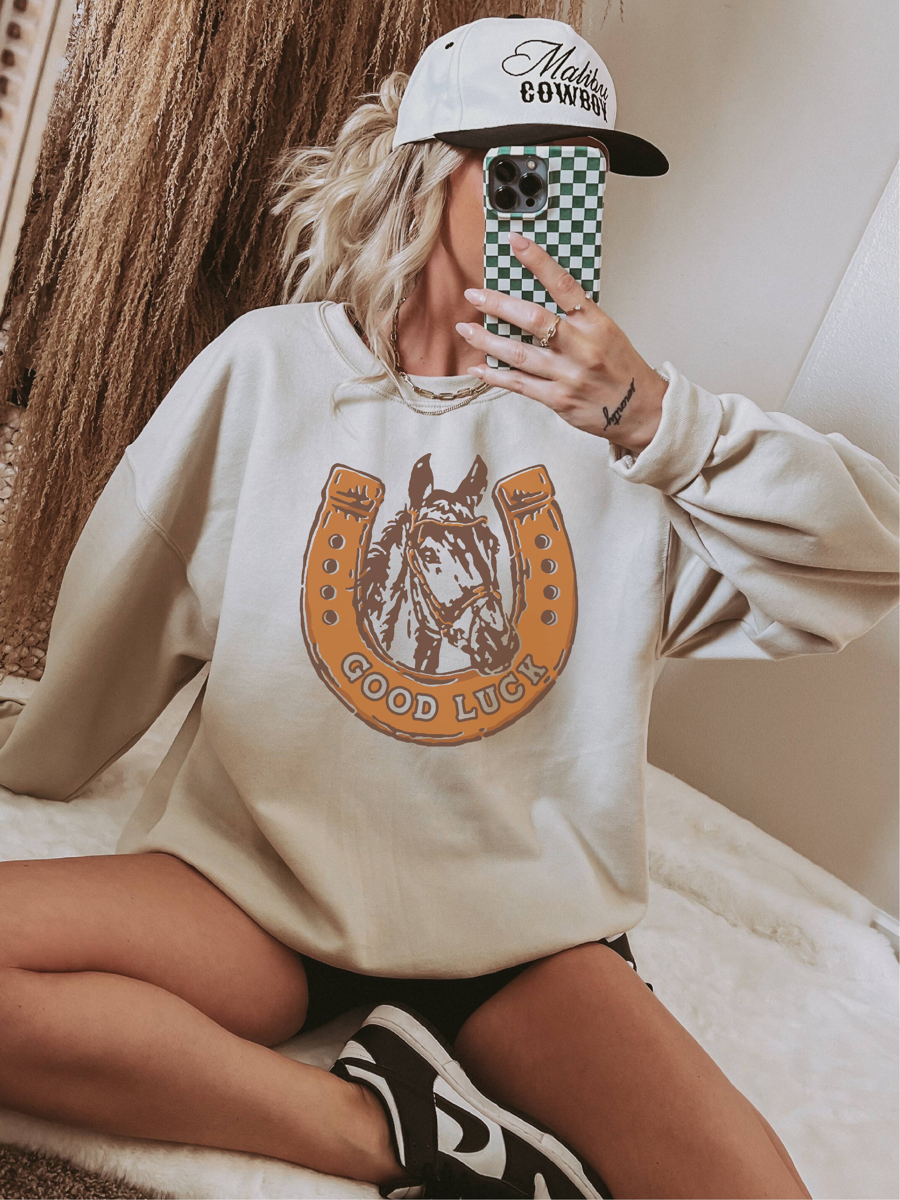 Person wearing a beige sweatshirt with a horse graphic and text, holding a phone with a checkered case.