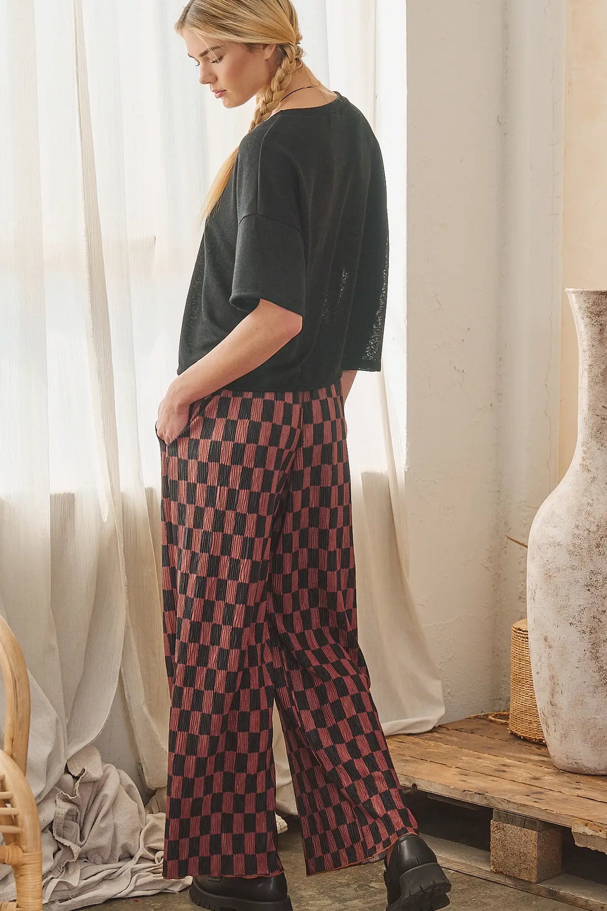 A woman stands in a bright room wearing loose black short-sleeve top and wide-leg checkered pants in red and black, with sheer curtains, a wooden bench, and a large vase in the background.