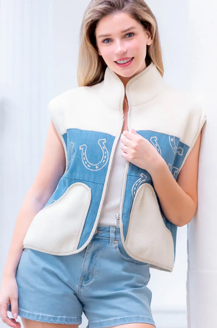 Model wearing indigo denim vest with white horseshoe print and faux-fur collar, zipped front, hands in pockets.”