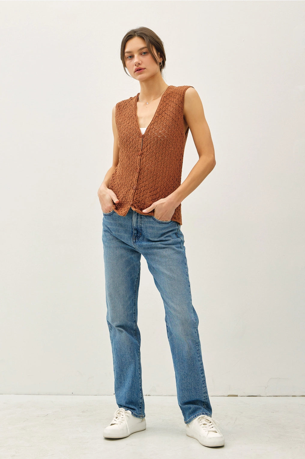 Woman wearing a brown knitted vest, blue jeans, and white sneakers on a white background