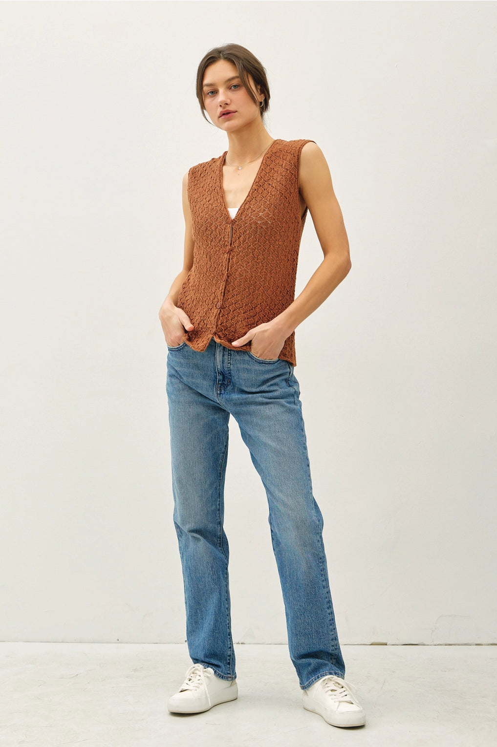 Woman wearing a brown knitted vest, blue jeans, and white sneakers on a white background