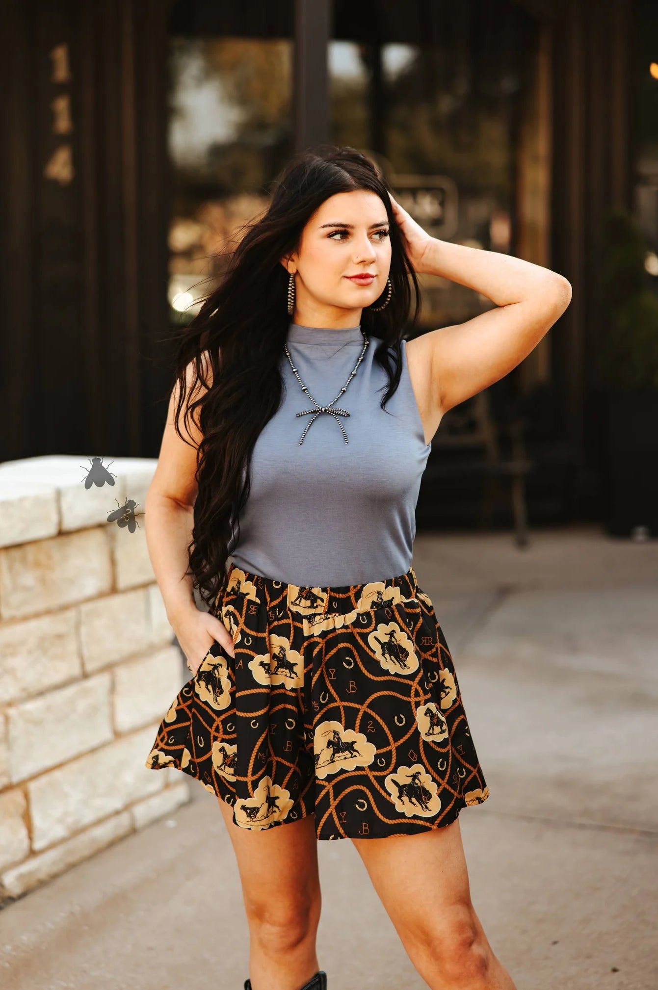 Woman wearing a steel blue top and patterned shorts standing outdoors.