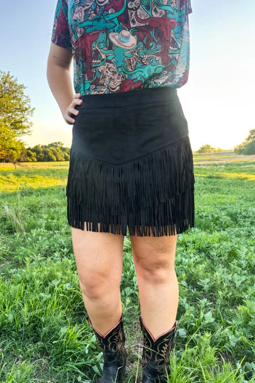 A person standing outdoors in a grassy field wearing a black fringe mini skirt, a colorful patterned top, and black cowboy boots.