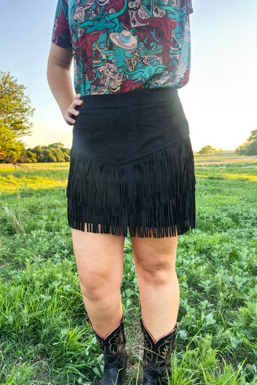 A person standing outdoors in a grassy field wearing a black fringe mini skirt, a colorful patterned top, and black cowboy boots.