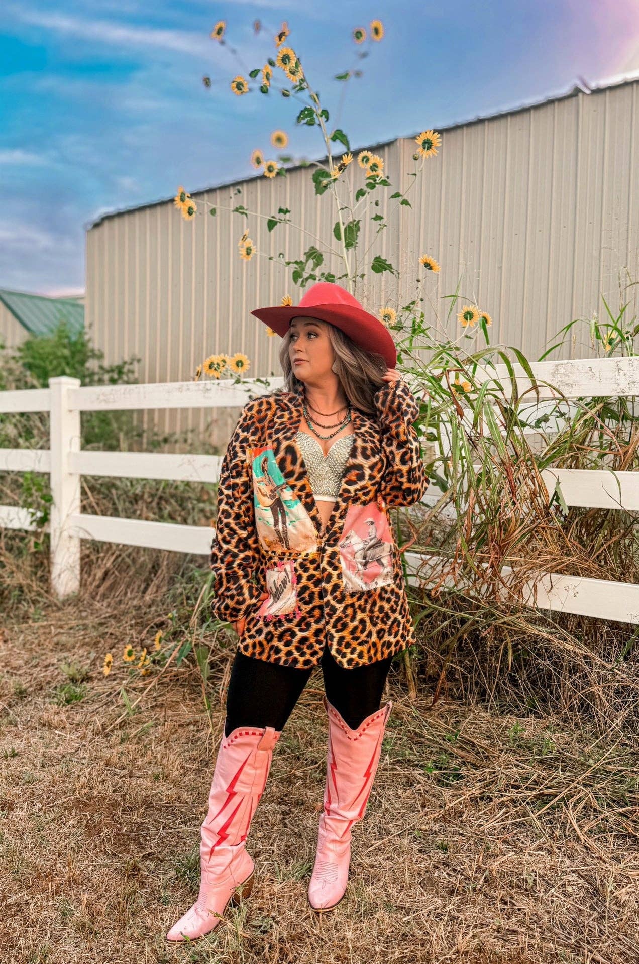 Person wearing a leopard print jacket and pink boots standing in a field with sunflowers and a barn in the background.