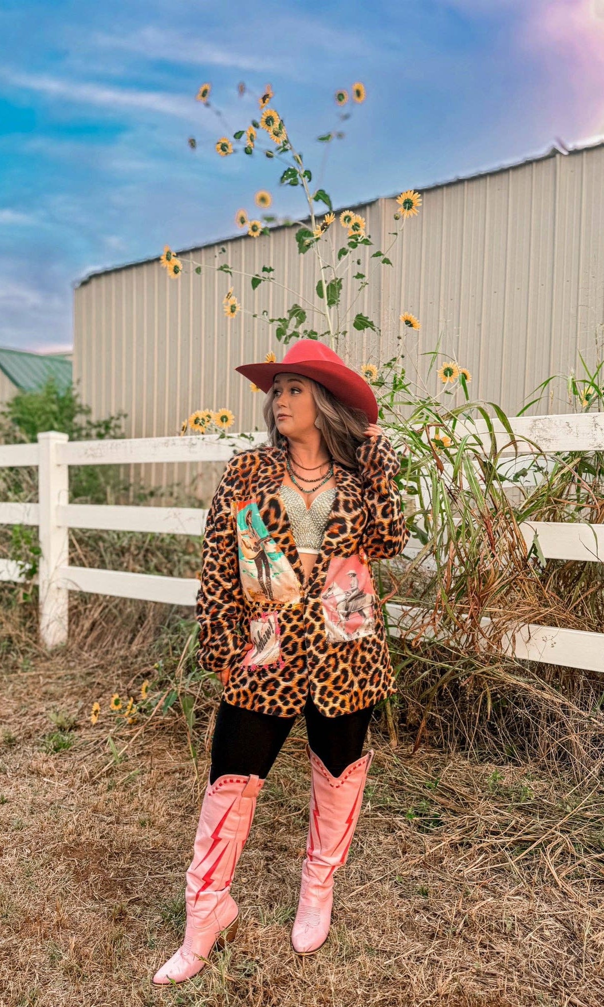 Person wearing a leopard print jacket and pink boots standing in a field with sunflowers and a barn in the background.
