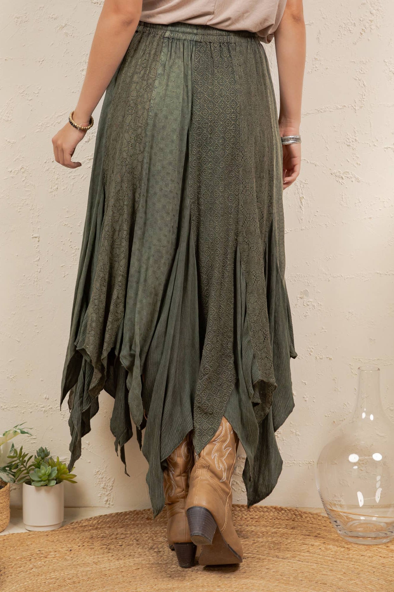 A woman wearing a flowing olive green high-low skirt with a subtle textured pattern, styled with tan cowboy boots in a rustic indoor setting.