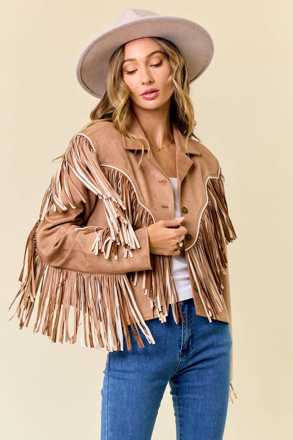 Woman wearing a tan fringe jacket, white top, and blue jeans against a beige background