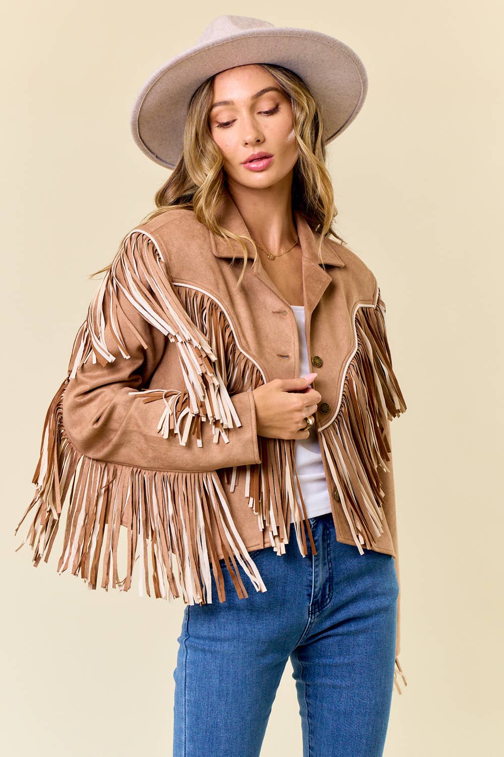 Woman wearing a tan fringe jacket, white top, and blue jeans against a beige background